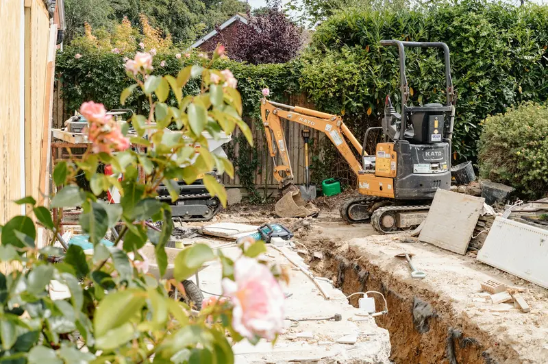 Mini excavator opening a trench along a garden wall beside a house