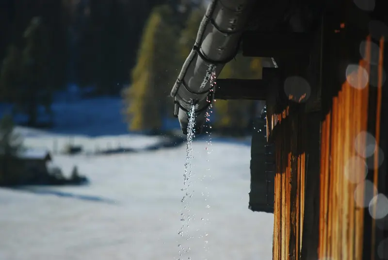 Downspout dripping meltwater beside a wooden wall in winter